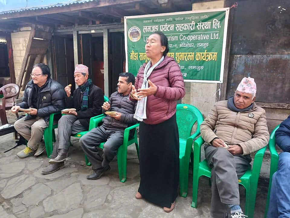 लमजुङ पर्यटन सहकारस् संस्था लिमिटेडको ब्यानरको अगाडी छुपिमाया गुरुङ मन्तब्य राख्दै हुनुहुन्छ भने उहाँको दायाँवाया पुरुषहरु कुर्सिमा बसिरहेका छन् । उनीहरु केही खाइरहेका छन् । 