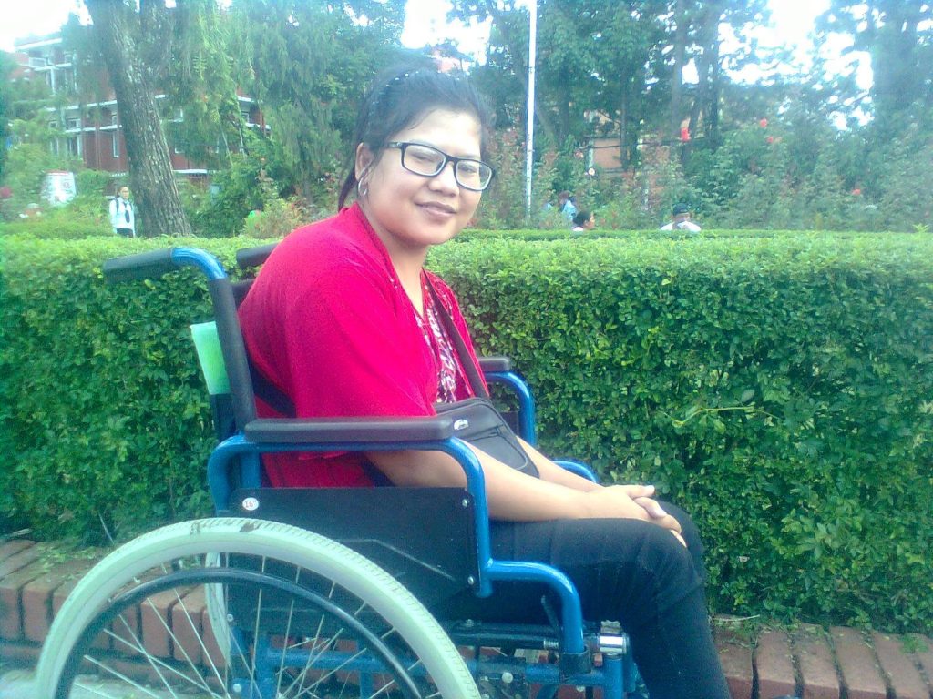 लेखक तथा सर्जक, अरुणा छन्त्याल ह्विल चेयरमा बसेर क्यामेरा तर्फ हेरी रहनु भएको छ । उहाँको फोटोको पछाडी बगैंचा रहेको छ । उहाँले चश्मा लगाउनु भएको छ । 